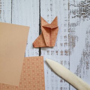 An origami fox on a pale wood background, with a folding tool and a couple of squares of origami paper